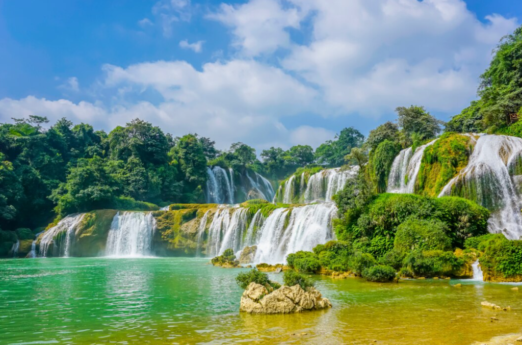 A breathtaking view of a large, multi-tiered waterfall cascading into a turquoise pool, surrounded by lush green vegetation under a bright blue sky, exemplifying staggering beauty.